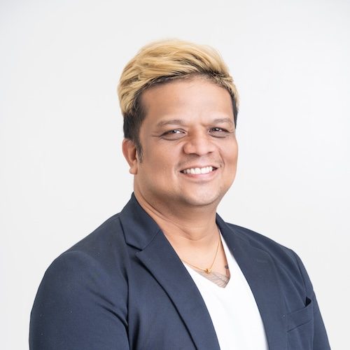 A person with short blonde hair, wearing a dark jacket over a white shirt, smiles in front of a plain white background, embodying the spirit of helping businesses go online.